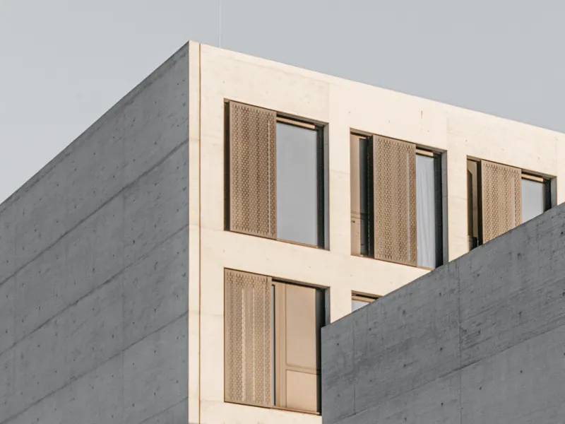 Façade de logements en béton clair et volets bois coulissants, vue en angle sous un ciel gris pâle.